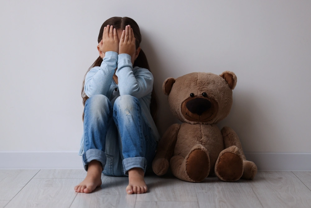 Upset little girl with teddy bear sitting on floor near light wall indoors
