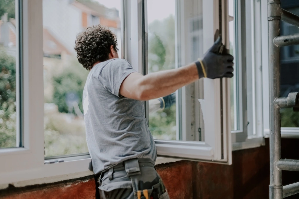 man in a gray uniform stands from the back and repairs installing a window frame in a house
