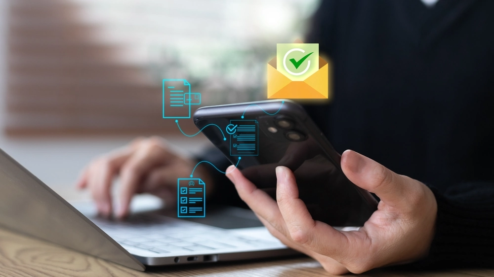 A hand using a smartphone and laptop with email and document approval icons