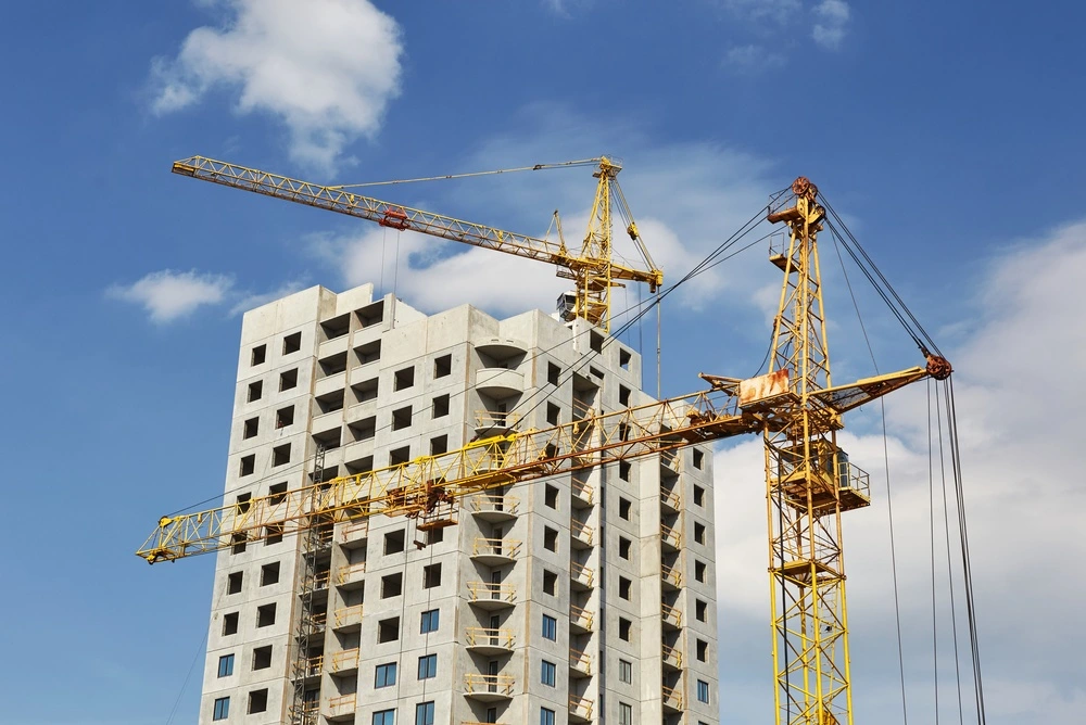 Two construction tower cranes operate against the backdrop of an unfinished multi-story building